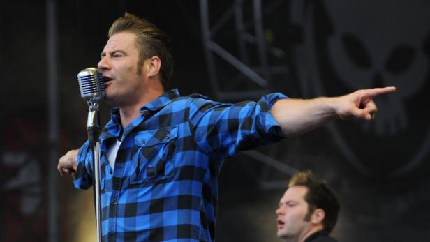 Ein Mann mit blau-schwarzem Hemd singt in ein Mikrofon und zeigt mit dem Finger.