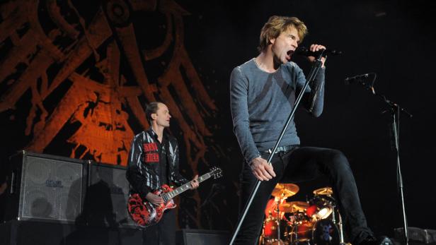 Ein Sänger performt mit Gitarrist und Schlagzeuger auf einer Bühne mit einem abstrakten Hintergrund.