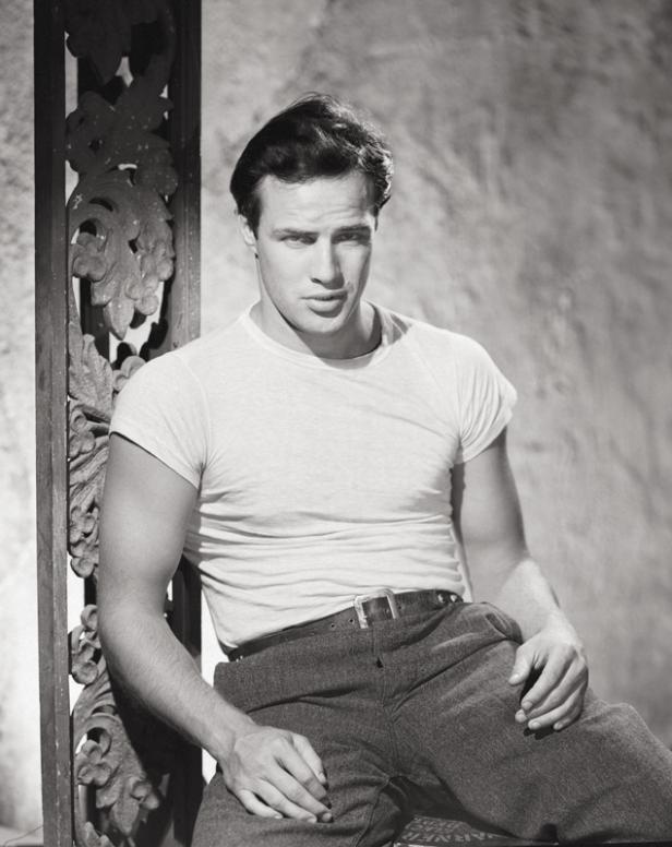 Marlon Brando sitzt in einem T-Shirt und einer Hose mit Gürtel vor einer Wand.