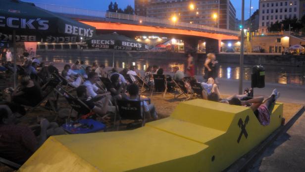 Menschen entspannen an einem Strandabschnitt am Wasser unter Sonnenschirmen der Marke Beck's.