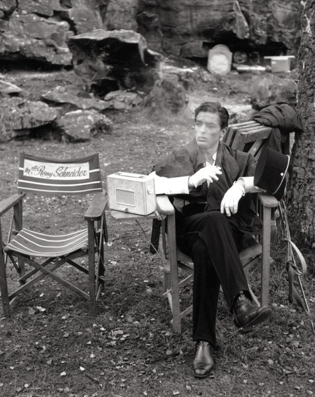 Ein Mann in formeller Kleidung sitzt rauchend in einem Regiestuhl mit der Aufschrift „m. Romy Schneider“.