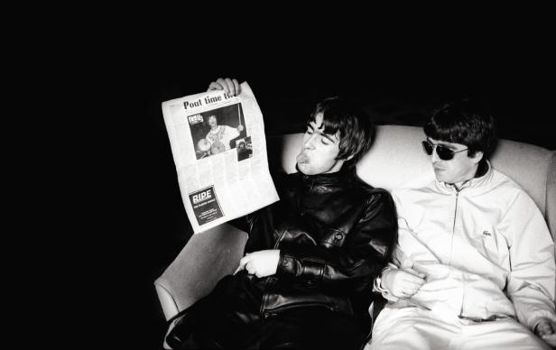 Liam und Noel Gallagher von Oasis sitzen auf einem Sofa, Liam liest Zeitung.