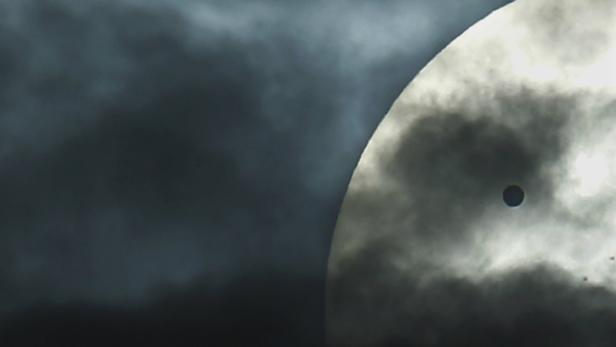 Ein dunkler Planet ist vor einer hellen Sonne zu sehen, teilweise von Wolken verdeckt.