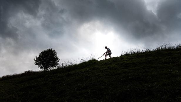 Eine Person mäht mit einer Sense einen steilen Hang unter einem bewölkten Himmel.