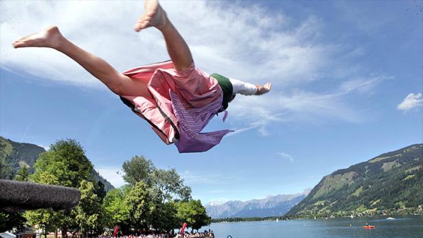 Ein Mann im Dirndl springt in einen See vor einer Bergkulisse.