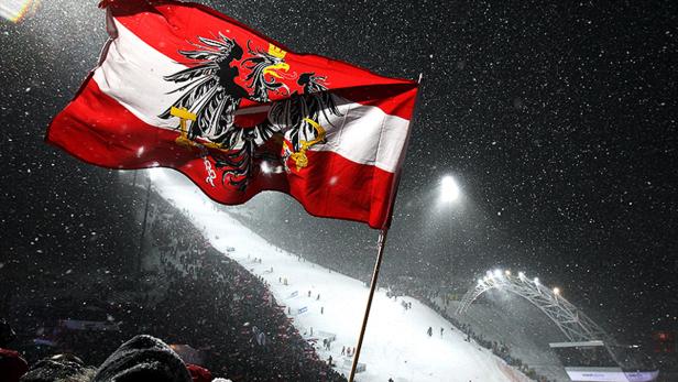 Eine österreichische Flagge weht vor einer beleuchteten Skipiste mit Zuschauern im Schnee.