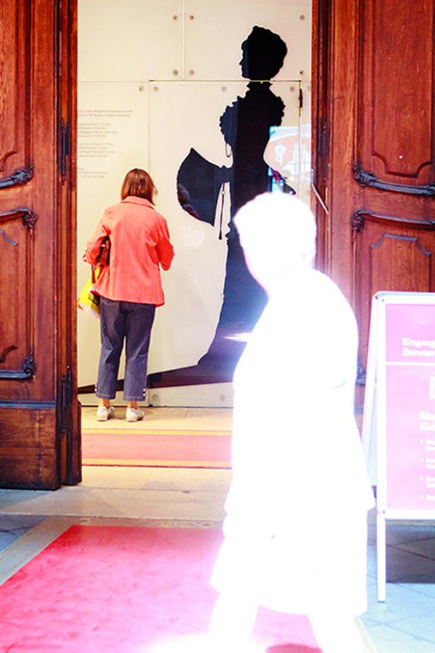 Besucher betreten eine Ausstellung mit einer Silhouette einer Frau im historischen Kleid.