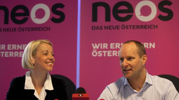 Eine Frau und ein Mann sitzen vor einem rosafarbenen Hintergrund mit dem NEOS-Logo.