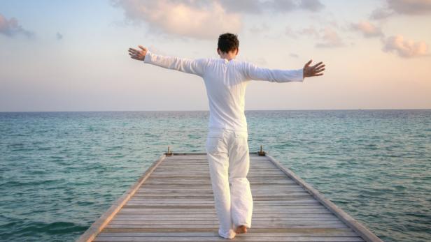 Ein Mann steht barfuß auf einem Holzsteg und blickt mit ausgestreckten Armen aufs Meer.