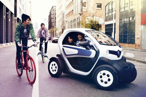 Zwei Radfahrer und ein Paar in einem Renault Twizy auf einer Straße in der Stadt.