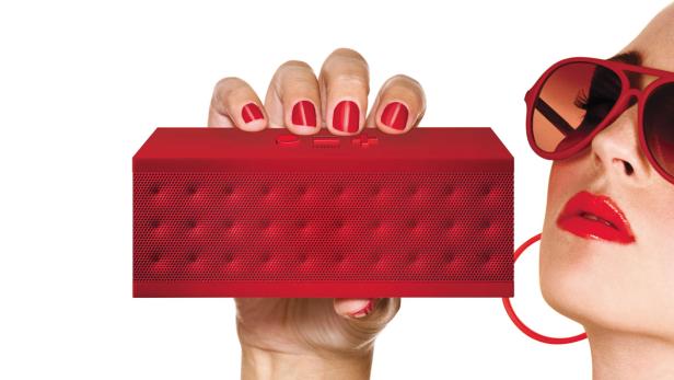Eine Frau mit rotem Lippenstift und Sonnenbrille hält einen roten JamBox-Lautsprecher.