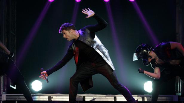 Ein Sänger performt auf einer Bühne mit Tänzern im Scheinwerferlicht.