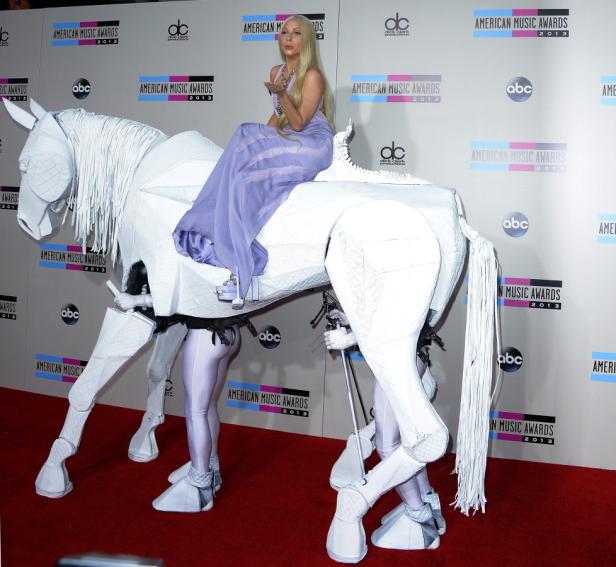 Lady Gaga reitet auf einem von zwei Personen dargestellten, weißen, mechanischen Pferd auf dem roten Teppich.