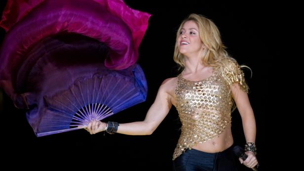 Shakira tritt in einem goldenen Oberteil mit einem lila Fächer auf einer Bühne auf.