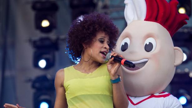 Eine Sängerin mit Afro-Frisur singt neben einem Maskottchen in den polnischen Nationalfarben.