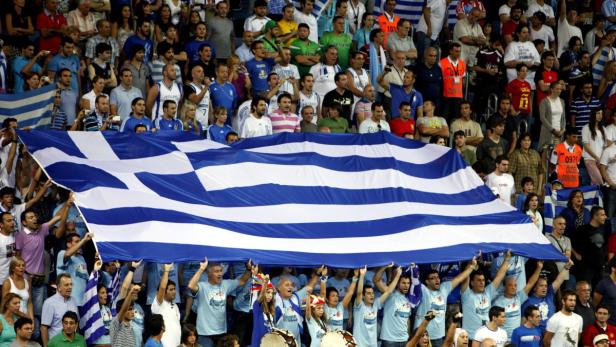 Eine Menschenmenge hält eine große griechische Flagge in einem Stadion hoch.