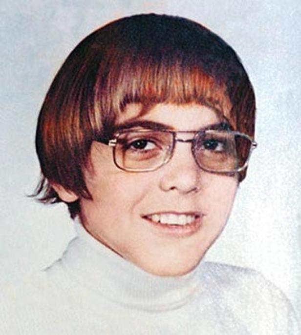 Ein junger George Clooney mit braunen Haaren, Pony und Brille lächelt.