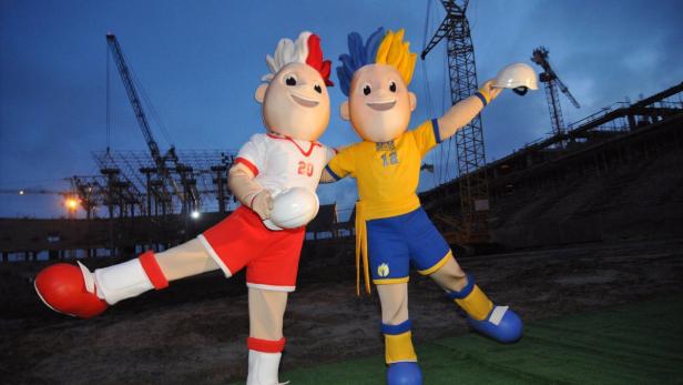 Zwei Maskottchen stehen vor einer im Bau befindlichen Arena.