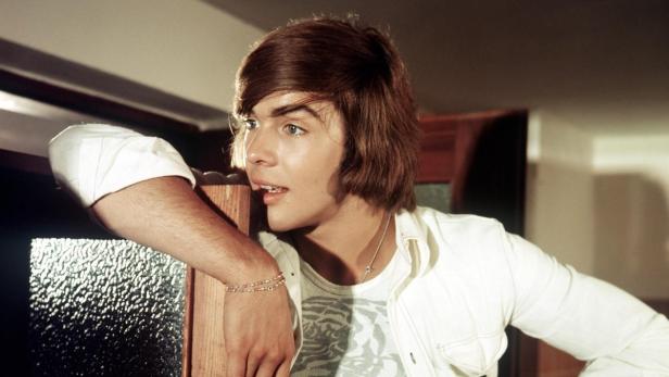 Ein junger Mann mit braunen Haaren und hellen Augen lehnt an einem Holzpfosten.