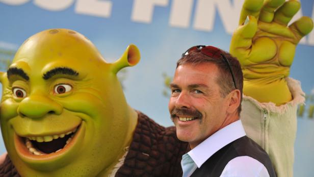 Ein Mann mit Schnurrbart posiert neben einer Shrek-Figur.