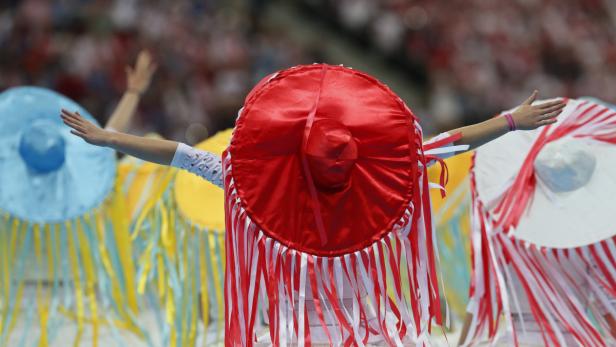 Tänzer tragen bunte, kreisförmige Kostüme mit Fransen und erhobenen Armen.