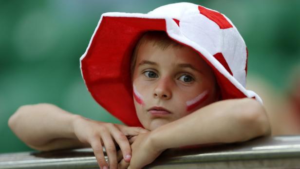 Ein Junge mit rot-weißem Hut und bemalten Wangen lehnt an einer Absperrung.