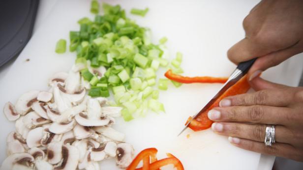 Eine Person schneidet Paprika auf einem Schneidebrett mit Pilzen und Frühlingszwiebeln.