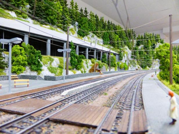 Eine Miniaturlandschaft mit einem Bahnhof und Gleisen vor einer bewaldeten Hügellandschaft.