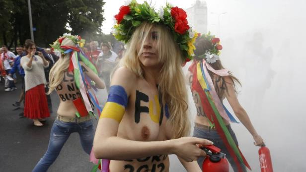 Drei barbusige Aktivistinnen mit Blumenkränzen protestieren mit Farbe und Feuerlöschern.