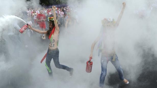 Zwei barbusige Frauen mit Blumenkränzen inmitten einer Rauchwolke, eine hält einen Feuerlöscher.