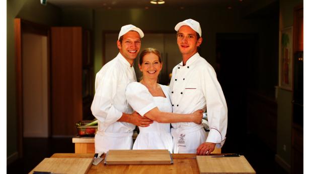 Eine Frau in Weiß posiert mit zwei Köchen in einer Küche.