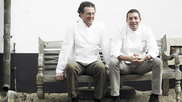 Zwei Köche in Kochjacken sitzen auf einer Holzbank vor einer weißen Wand.