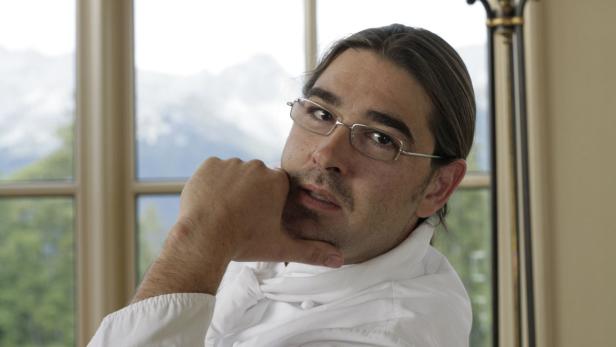 Ein Mann mit Brille und langem Haar posiert vor einem Fenster mit Bergblick.