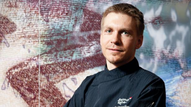 Ein Mann in Kochkleidung steht vor einem abstrakten Hintergrund.