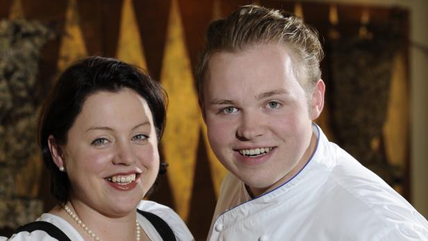 Ein Mann in Kochuniform und eine Frau mit Perlenkette lächeln in die Kamera.