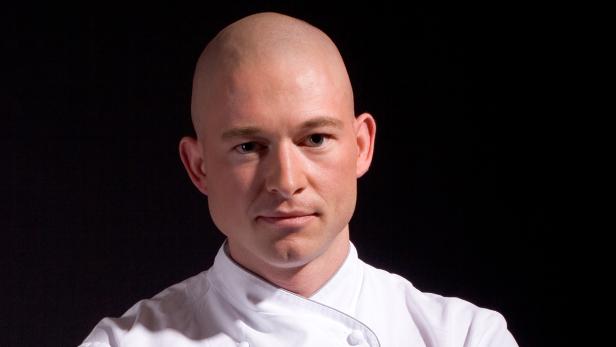 Ein Mann mit rasiertem Kopf und Kochjacke vor schwarzem Hintergrund.