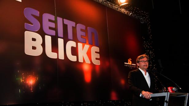 Ein Mann spricht vor einer Wand mit der Aufschrift „Seitenblicke“.