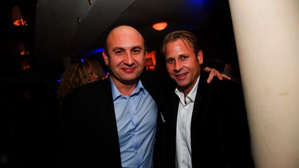 Zwei Männer in Anzügen posieren für ein Foto in einem dunklen Raum.