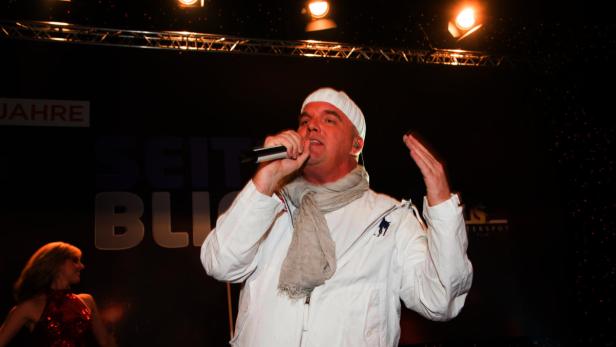 Ein Mann mit Mütze und Schal singt auf einer Bühne in ein Mikrofon.