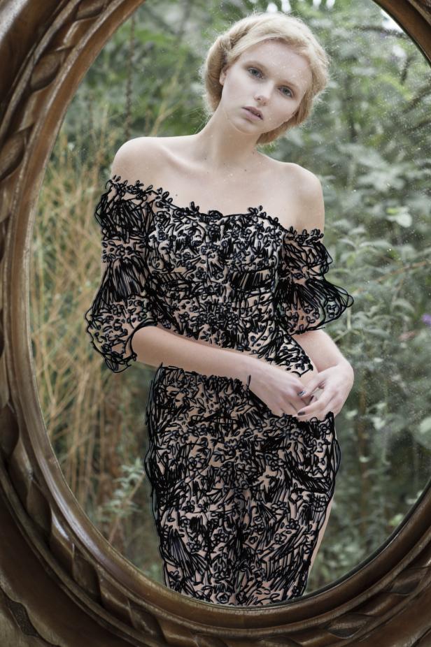 Eine junge Frau mit blonden Haaren, gesehen durch einen ovalen Spiegel, trägt ein schwarzes Kleid.