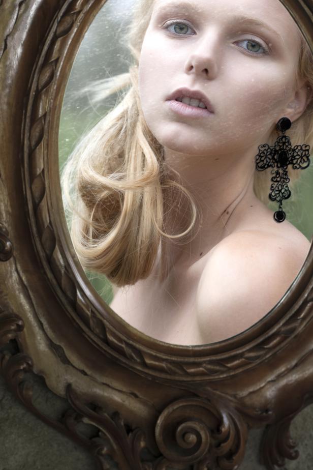 Eine junge Frau mit blonden Haaren und einem Ohrring blickt in einen verzierten Spiegel.