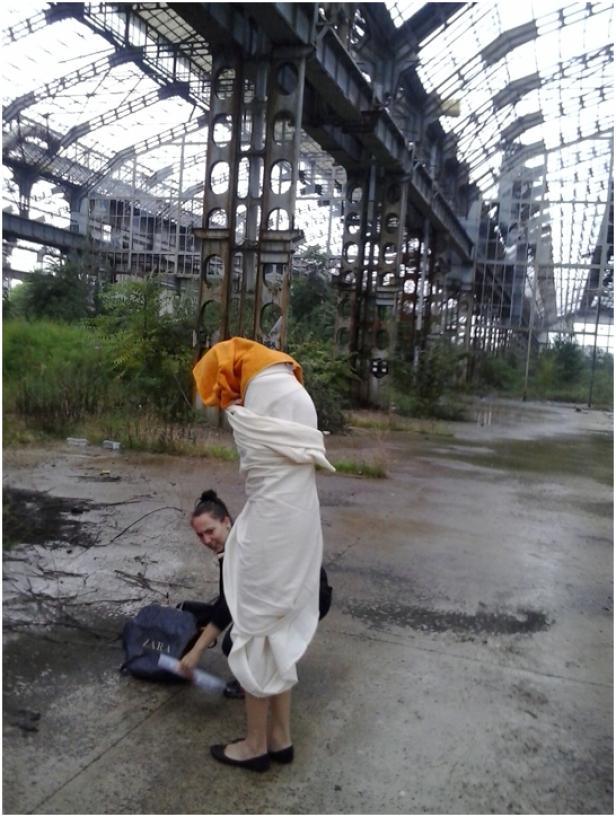 Eine Frau in einem weißen Kostüm posiert vor einer verlassenen Industriehalle, während eine andere Frau eine Tasche packt.