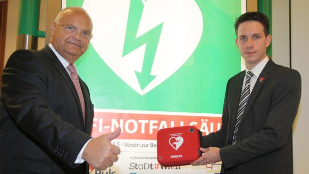 Zwei Männer in Anzügen präsentieren einen Defibrillator vor einem Schild.