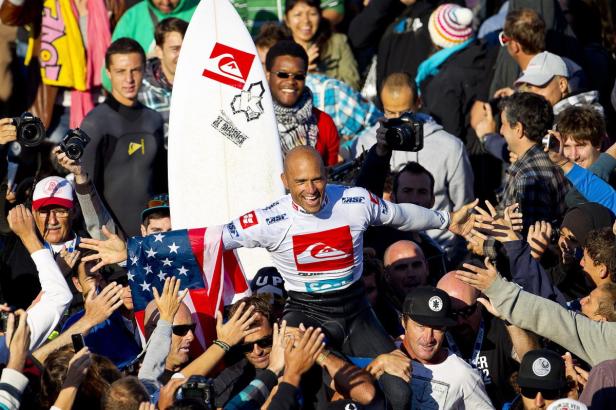 Kelly Slater wird nach einem Surfwettbewerb auf den Schultern getragen und hält eine US-Flagge.