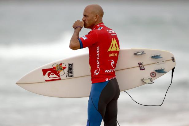Ein Surfer mit rotem Shirt und Surfbrett am Strand, die Hand am Mund.
