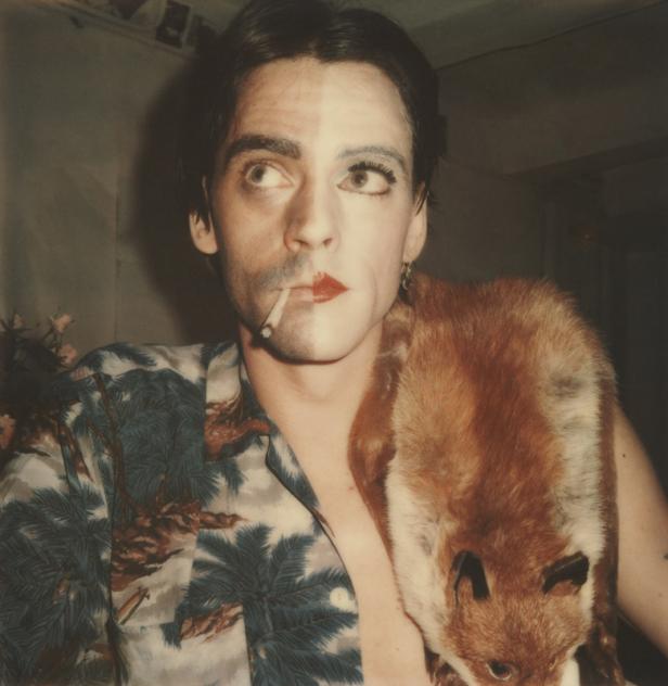 Ein Mann mit Make-up und einem Fuchsfell über der Schulter raucht eine Zigarette.