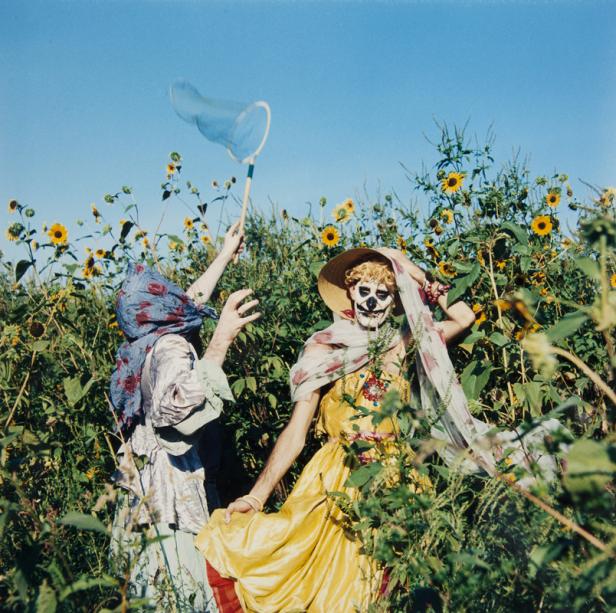 Zwei Personen in Kostümen und mit Gesichtsbemalung stehen in einem Feld mit Sonnenblumen.