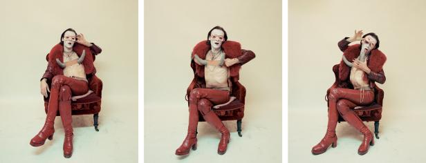 Eine Person mit Maske, Hörnern und roter Kleidung sitzt auf einem Sessel.