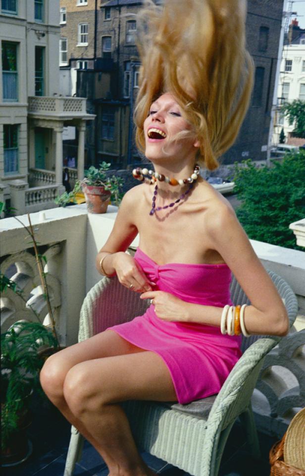 Eine Frau sitzt auf einem Balkon, wirft ihr blondes Haar zurück und lacht.