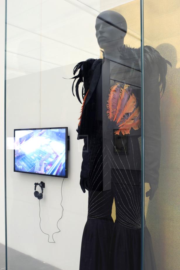 Eine Schaufensterpuppe trägt ein schwarzes Outfit mit Federn und ein Bildschirm zeigt ein Video.